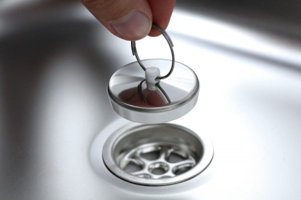 Ein Hand, der einen Abflussdeckel aus Metall über einem Waschbecken hält, ideal für mobile Wasserversorgungslösungen beim Camping oder im Transporter.