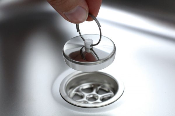 Ein Hand, die einen Abflussdeckel aus Edelstahl anhebt, um den darunterliegenden Abfluss in einer mobilen Waschstation zu zeigen. Ideal für Campingbedarf und mobile Wasserversorgungslösungen, wie Waschbecken und Wasserkanister.
