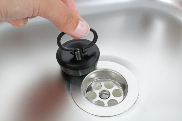 Hand, die einen schwarzen Abflussstopfen über einem Edelstahlwaschbecken hält, ideal für mobile Wasserversorgung beim Camping oder im Transporter.